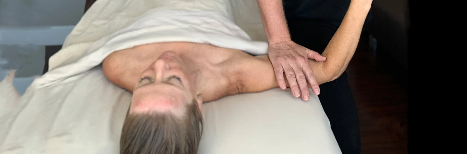 A massage therapist in black clothing performs deep tissue massage on a client's arm. The client lies face-up on a massage table with white linens, eyes closed in a relaxed state.