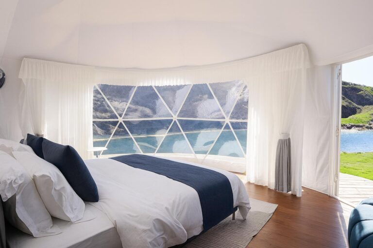 Interior of a geodesic dome bedroom featuring white bed with navy blue accent pillows, triangular window panels revealing lakeside landscape.
